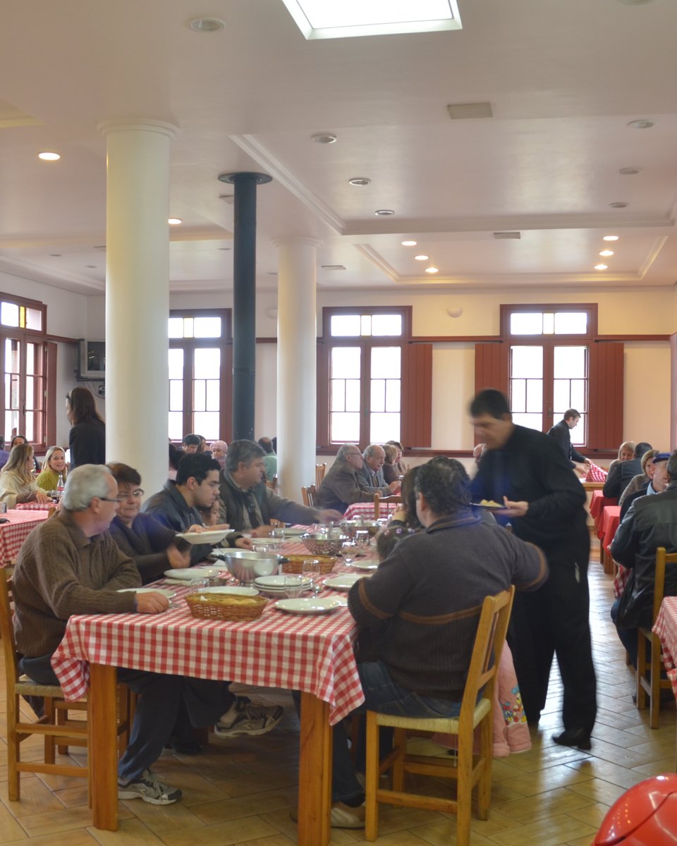 Salão de uma cantina no Vale dos Vinhedos, na Serra Gaúcha, com mesas de toalha xadrez vermelha, visitantes almoçando em família, um garçom servindo e janelas altas de madeira ao fundo.