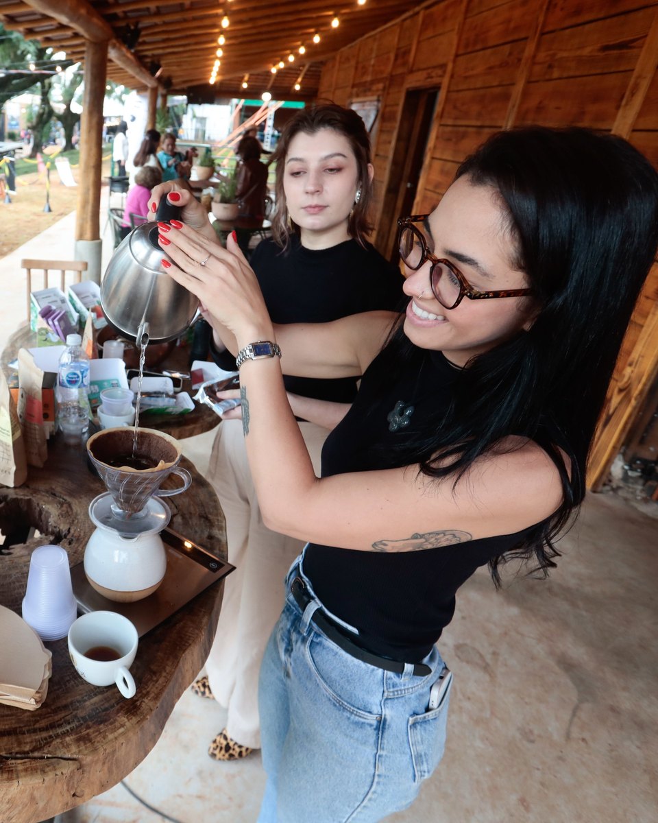 Mulher de óculos servindo café coado com uma chaleira de bico fino sobre uma mesa de tronco, num espaço de madeira com varal de luzes, no parque da Expoingá, em Maringá.