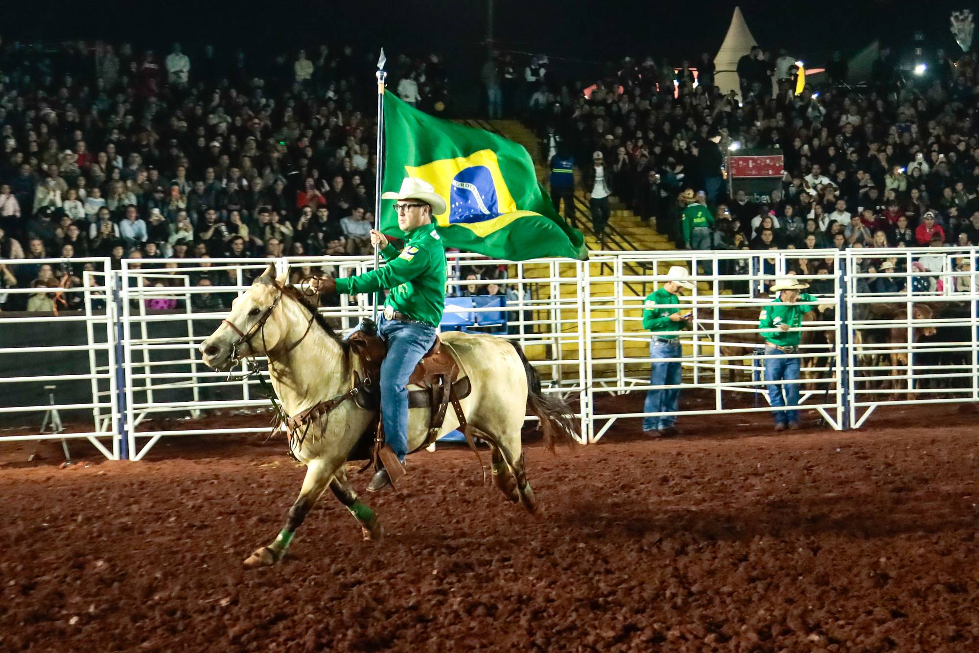 Cavaleiro de chapéu e camisa verde galopando com a bandeira do Brasil na arena de terra do rodeio da Expoingá, em Maringá, diante de uma arquibancada lotada à noite.