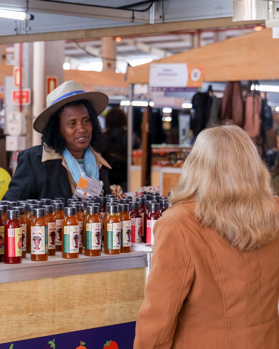 Produtora de chapéu de palha e lenço azul atendendo uma cliente de casaco caramelo numa banca de molhos de pimenta no Pavilhão da Agricultura Familiar da Expointer, em Esteio.