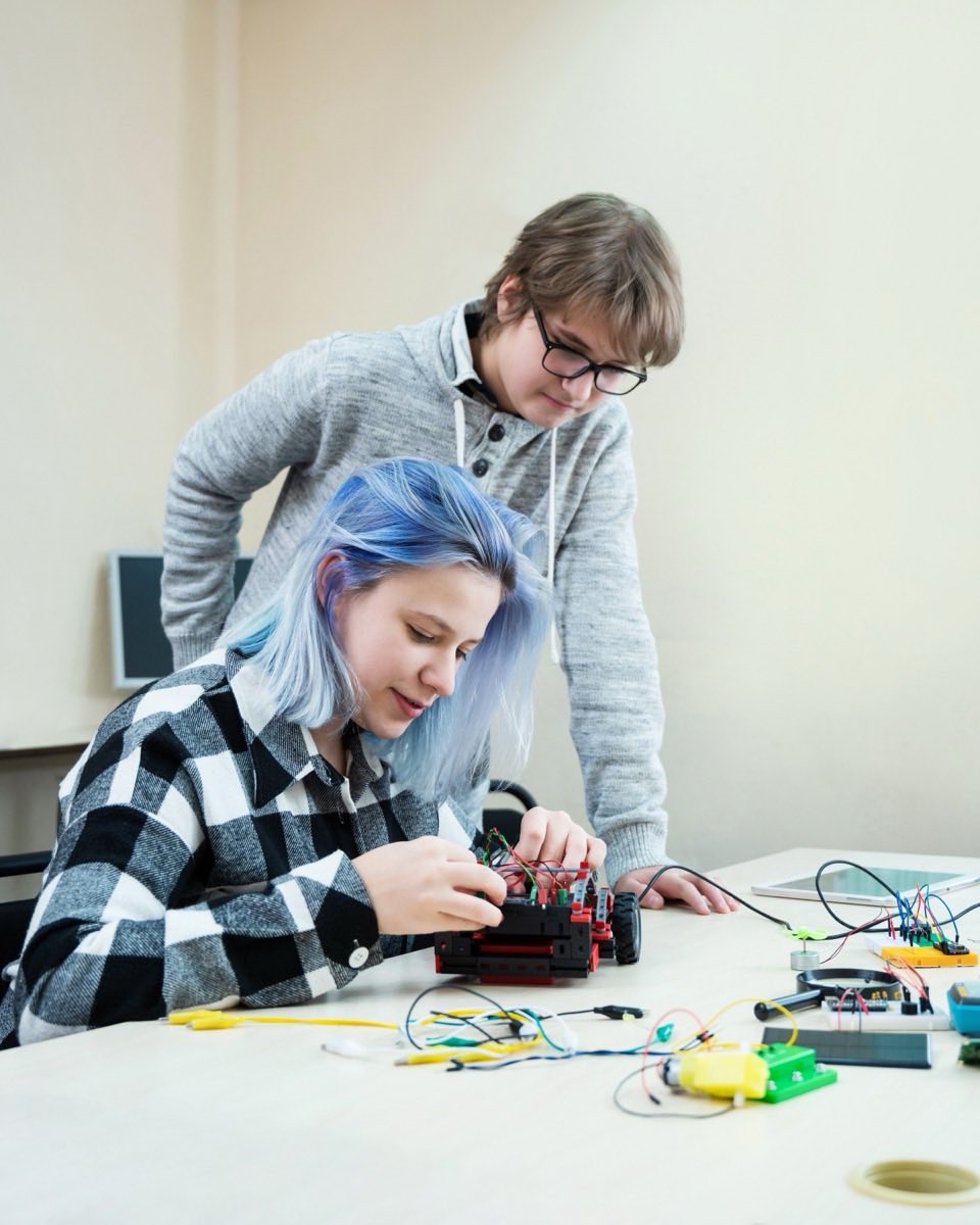 Dois estudantes montando um carrinho robô sobre uma mesa cheia de fios coloridos e placas eletrônicas, numa sala de aula de robótica.