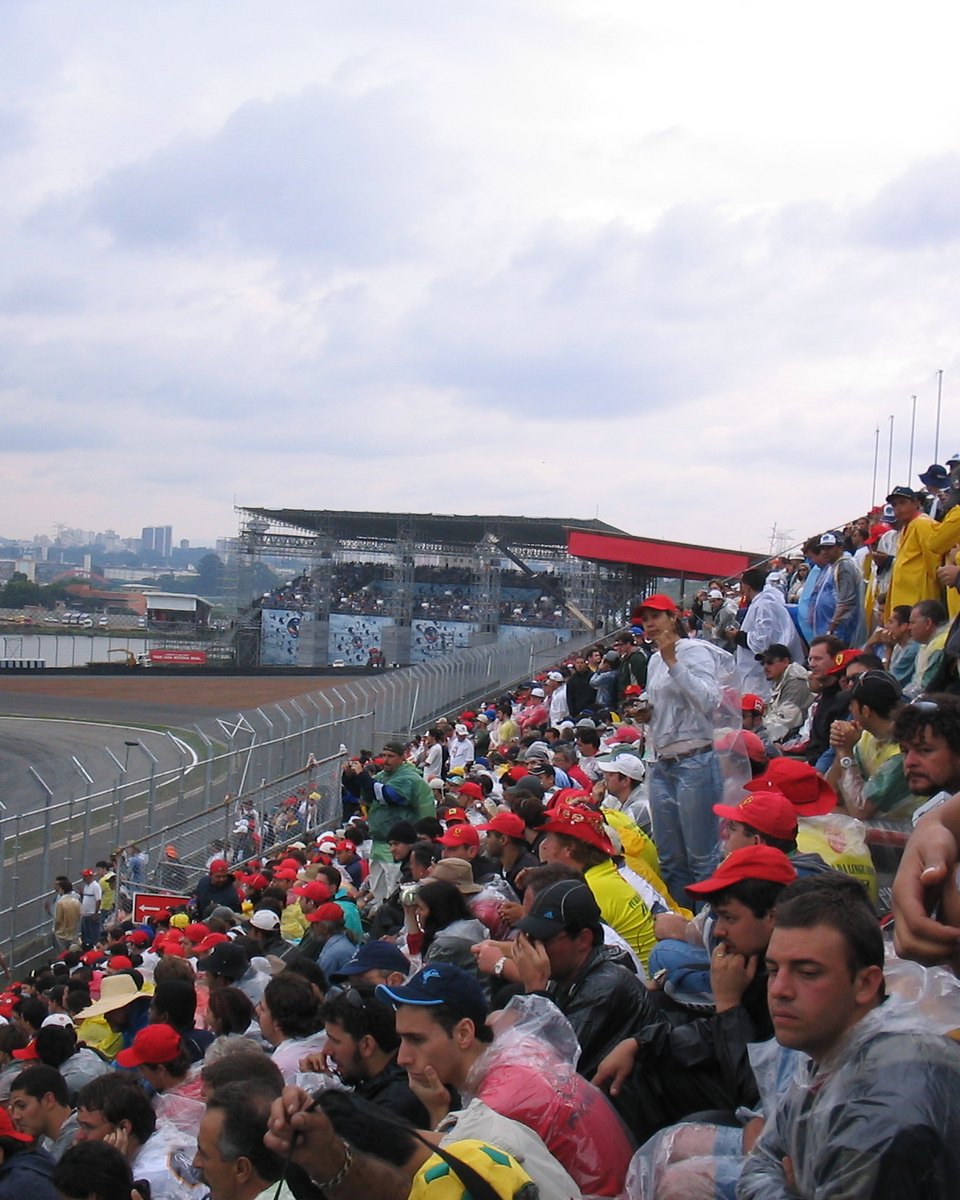 Arquibancada lotada de torcedores de boné em Interlagos, com a pista e os boxes ao fundo, em São Paulo.