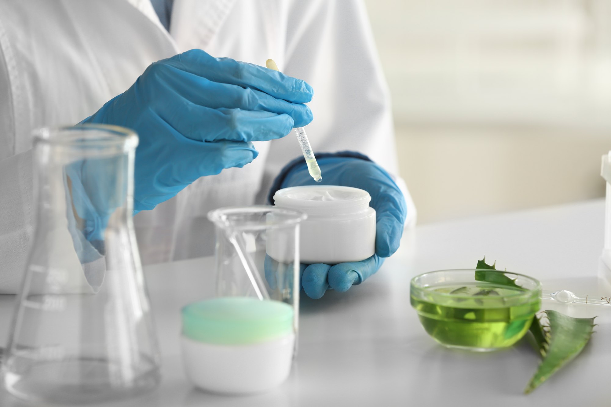 Mãos de luva azul pingando um ativo com conta-gotas num pote de creme branco, numa bancada de laboratório com béqueres e uma tigela de gel de babosa.