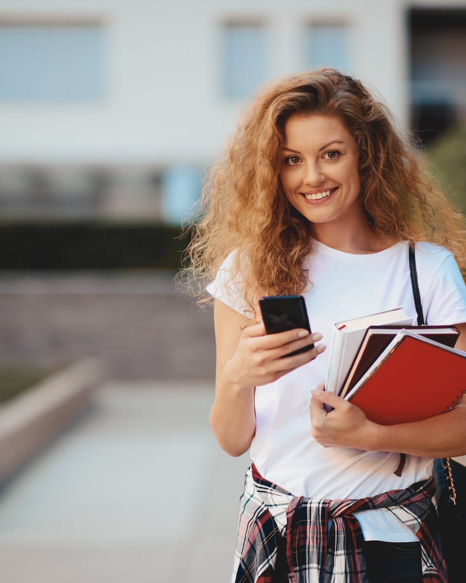 Estudante de cabelo cacheado e camiseta branca sorrindo com o celular na mão e cadernos e livros de capa vermelha junto ao corpo, caminhando por uma calçada arborizada de campus.