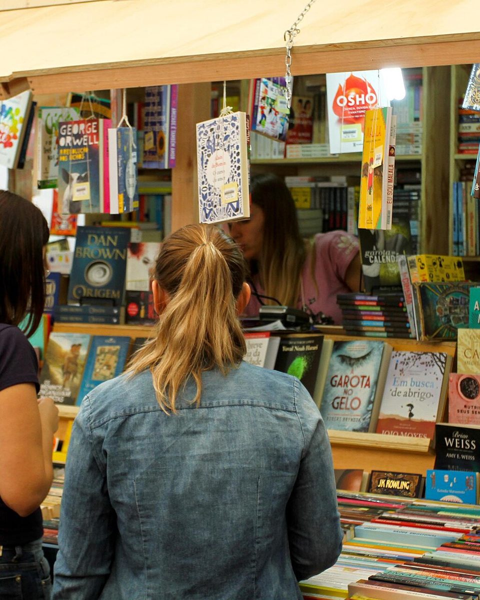 Duas mulheres de costas, uma de camiseta azul-marinho e outra de jaqueta jeans, escolhendo livros numa banca de madeira da Feira do Livro na Praça da Alfândega, em Porto Alegre, com livros pendurados e empilhados até o teto.