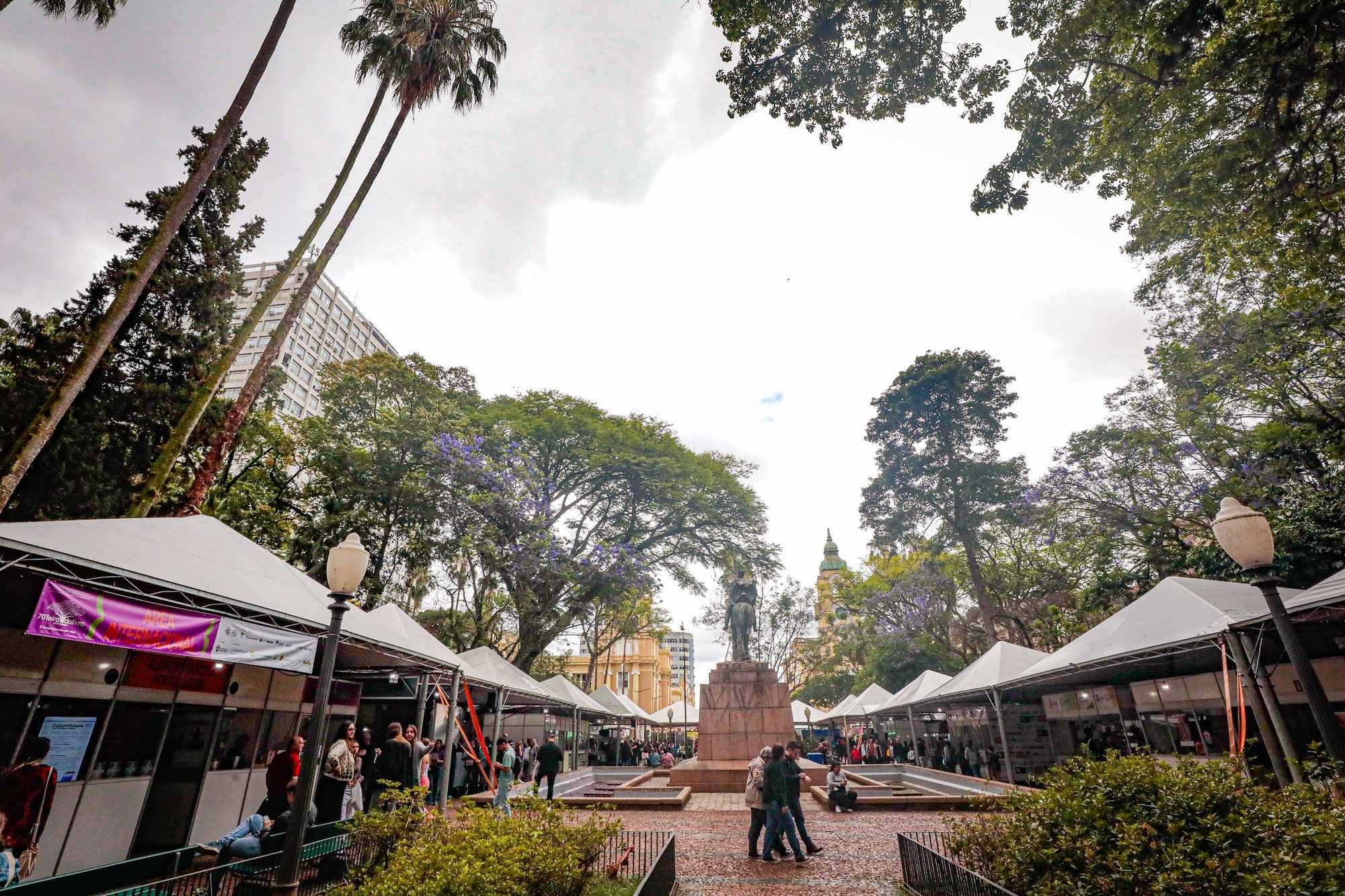 Bancas brancas da Feira do Livro enfileiradas dos dois lados da Praça da Alfândega, em Porto Alegre, com jacarandás de flores lilás, palmeiras, visitantes caminhando e um monumento com estátua equestre ao centro.
