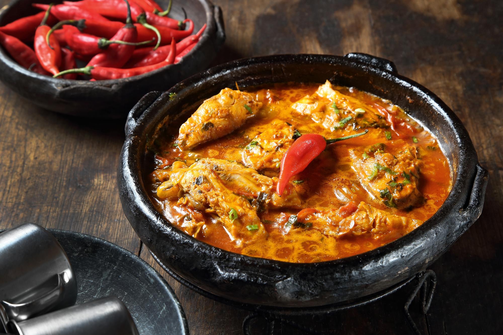 Moqueca capixaba de peixe fervendo numa panela de barro preta sobre mesa de madeira, com pimentas vermelhas numa tigela de barro ao lado.
