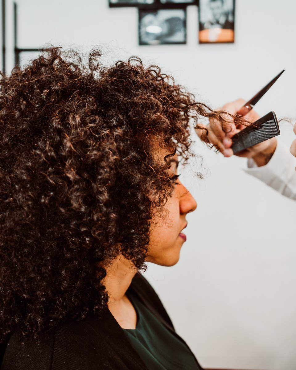 Cliente de cabelo cacheado castanho sentada num salão de beleza enquanto o cabeleireiro corta e modela os cachos com pente e tesoura.