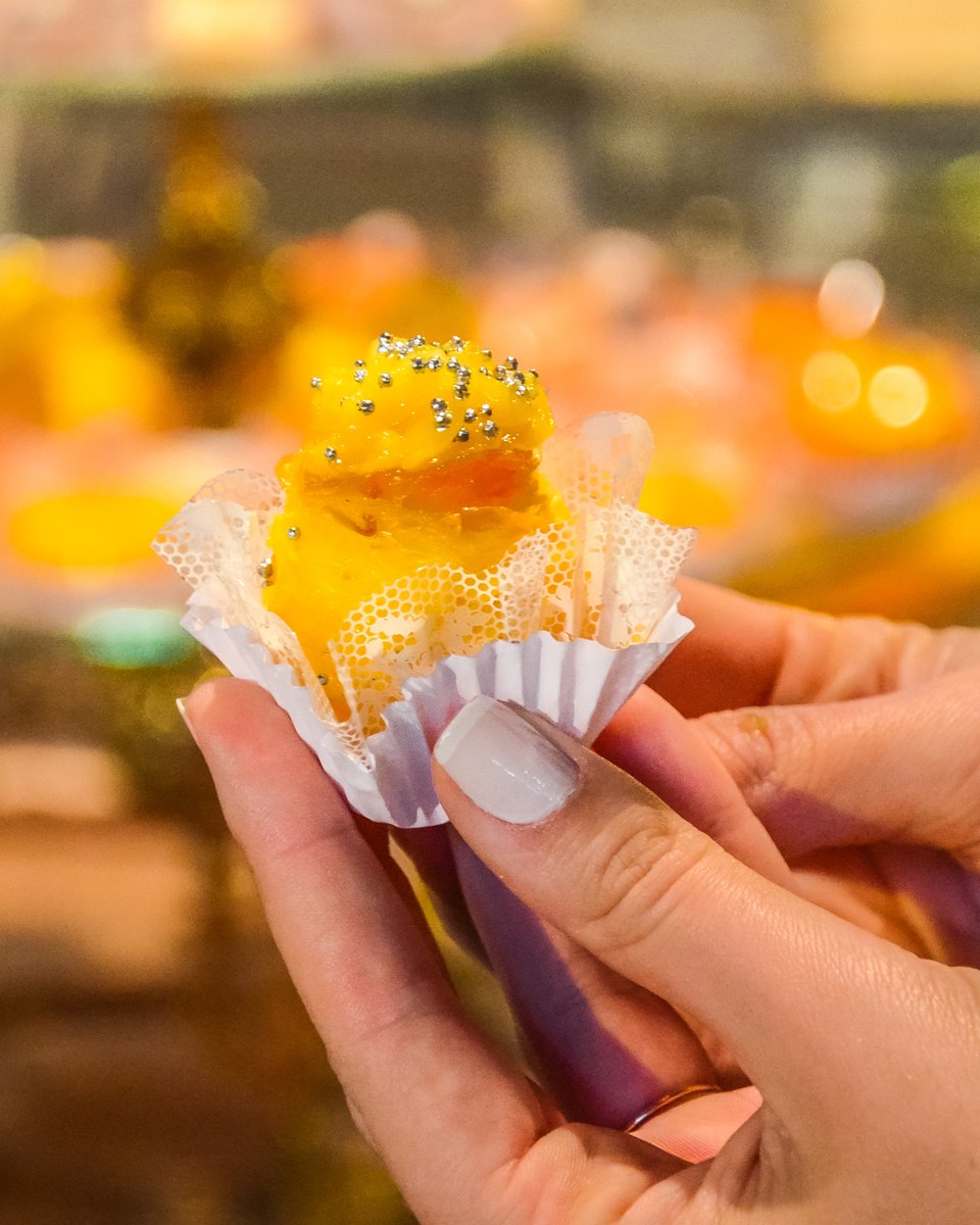 Mão de uma visitante segurando um doce fino amarelo, com confeitos prateados, numa forminha de papel rendada, diante de uma vitrine de doces desfocada, na Fenadoce, em Pelotas.