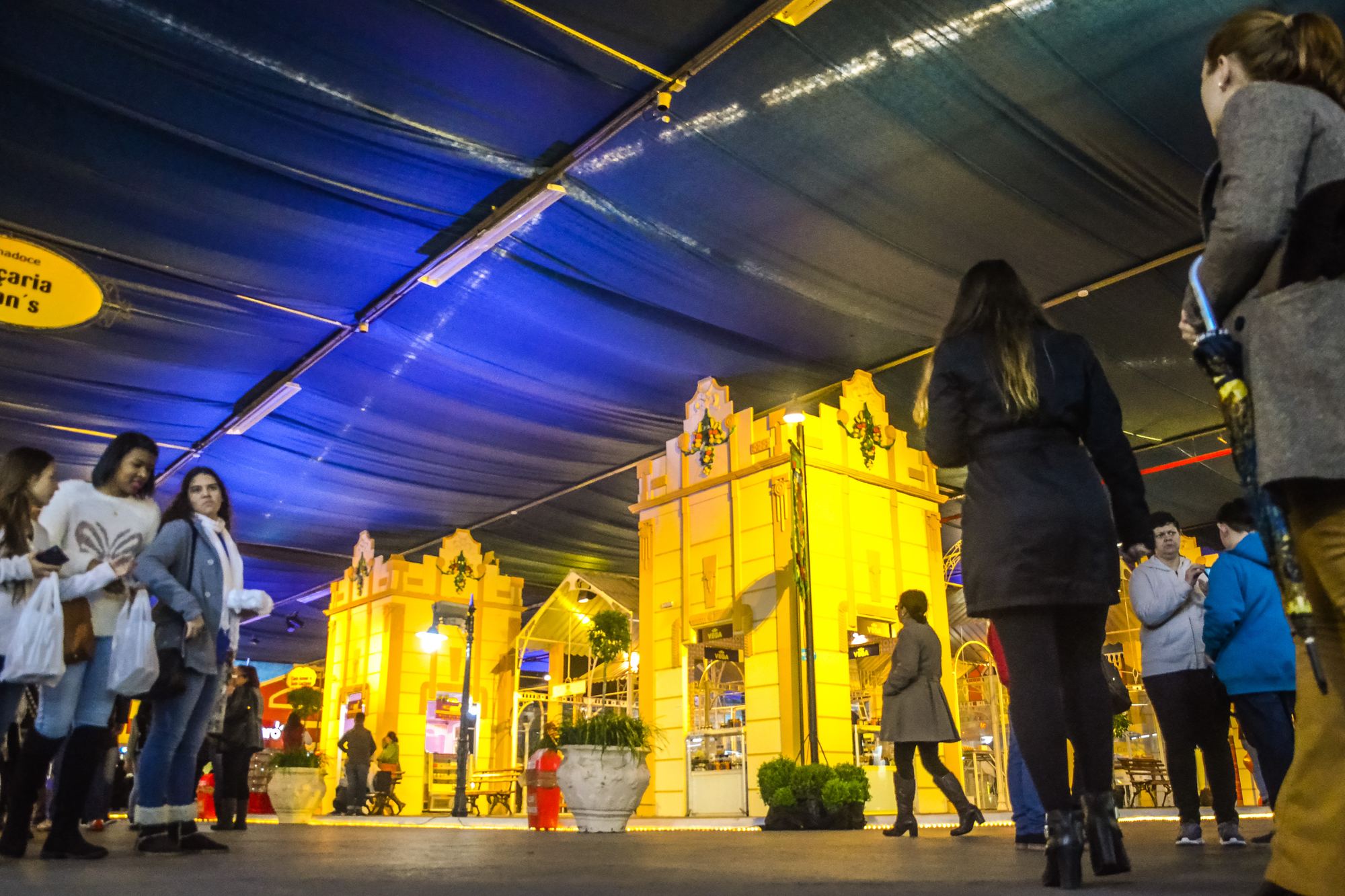 Visitantes de casaco caminhando pela Cidade do Doce, na Fenadoce, em Pelotas, entre fachadas amarelas iluminadas que imitam o casario antigo da cidade, sob a cobertura azul do pavilhão, com sacolas de doces na mão.