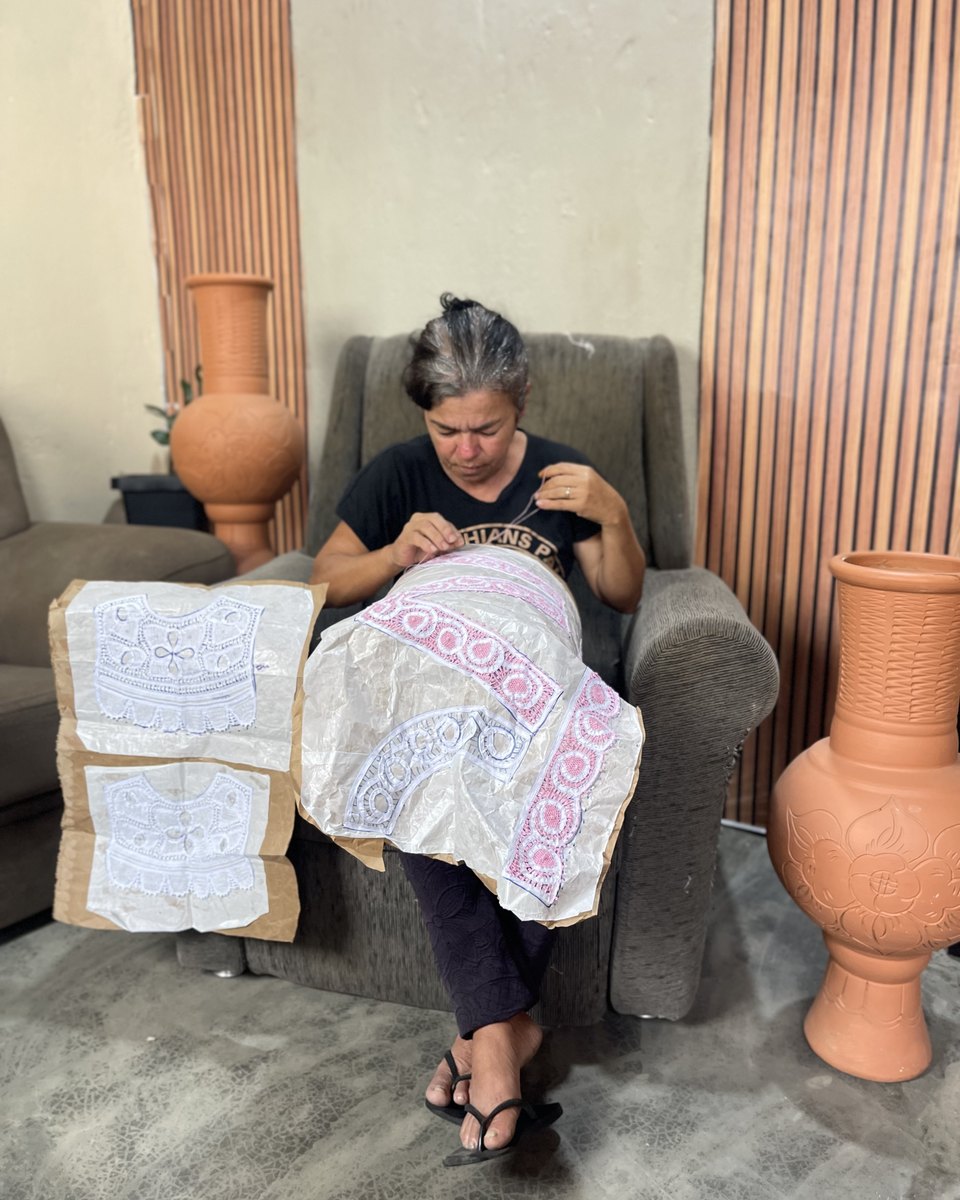 Rendeira sentada numa poltrona tece renda renascença branca e rosa sobre um molde de papel apoiado no colo, com riscos de renda ao lado e grandes vasos de barro na sala.