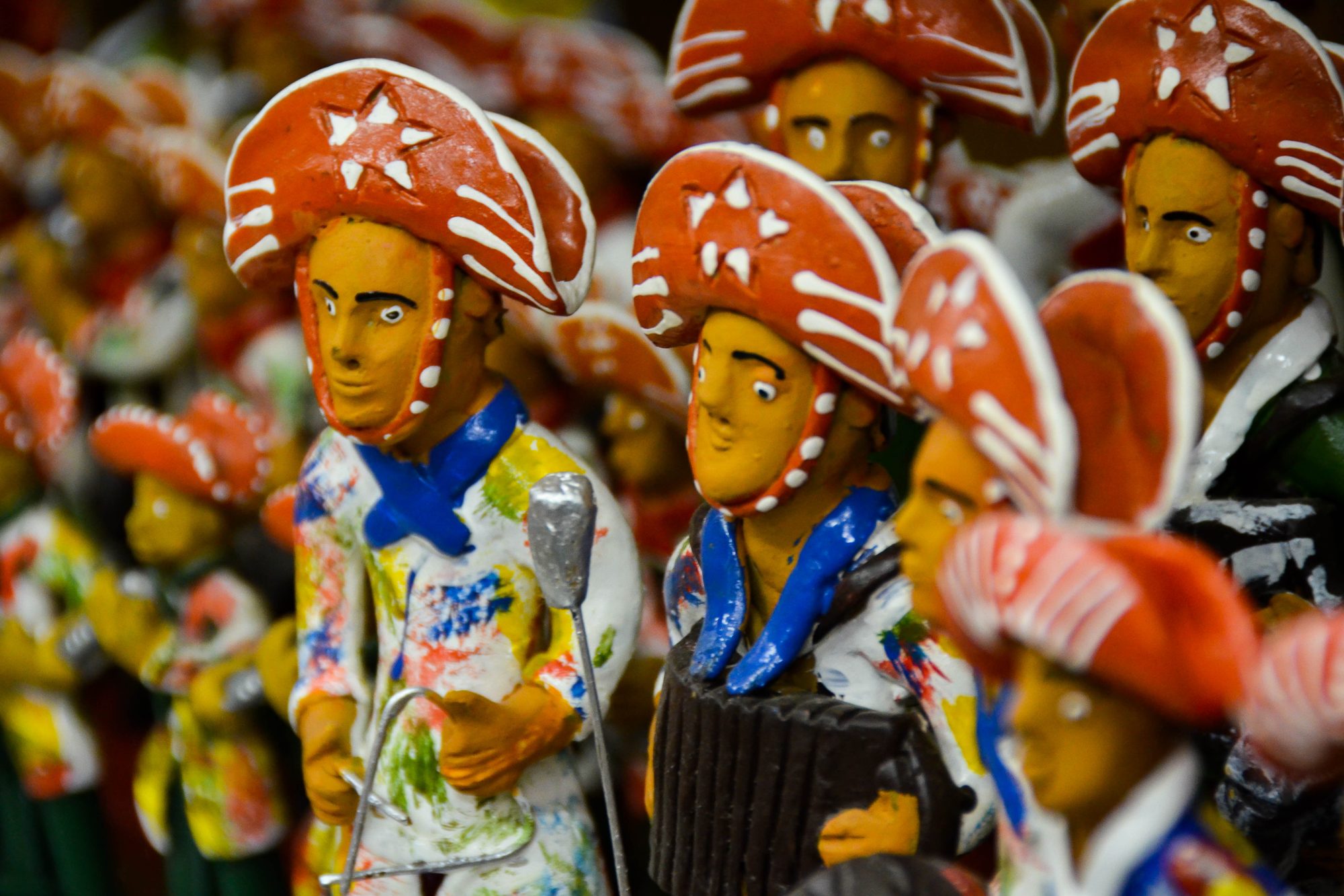 Bonecos de barro pintados de Caruaru, músicos de forró com chapéu de couro vermelho enfeitado de estrelas brancas, lenço azul, sanfona e triângulo, lado a lado numa prateleira.