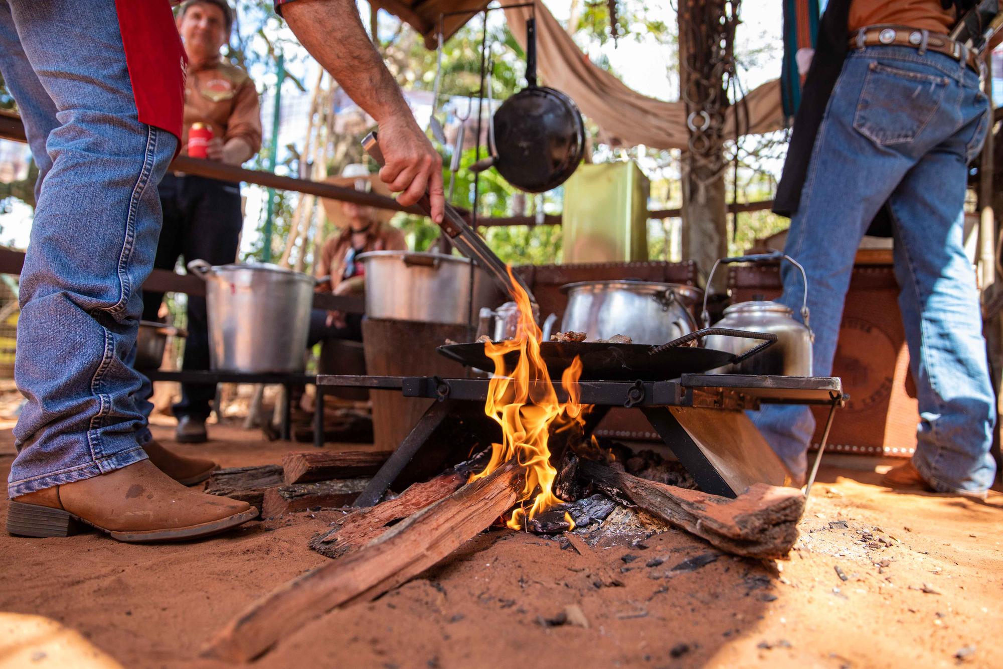 Homens de calça jeans e bota de couro em volta de uma fogueira no chão de terra vermelha, com panelas de ferro e alumínio, na Queima do Alho da Festa do Peão de Barretos.