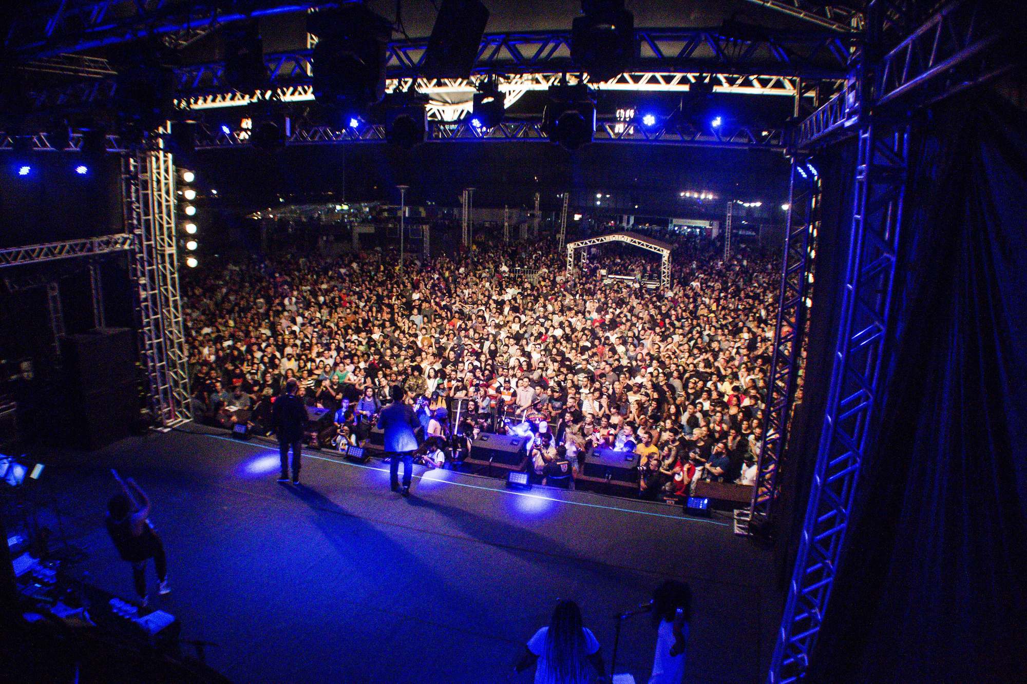 Vista de cima do palco para uma multidão que ocupa toda a arena do Festival Bananada, em Goiânia, à noite, com estrutura de treliças e luzes azuis em volta e músicos de costas no palco.