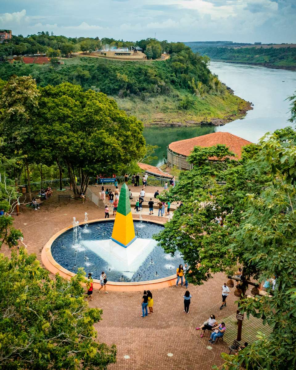 Vista do alto do Marco das Três Fronteiras, em Foz do Iguaçu: obelisco verde e amarelo no meio de um chafariz, turistas caminhando e sentados na praça e, ao fundo, o encontro dos rios Iguaçu e Paraná com a margem argentina.
