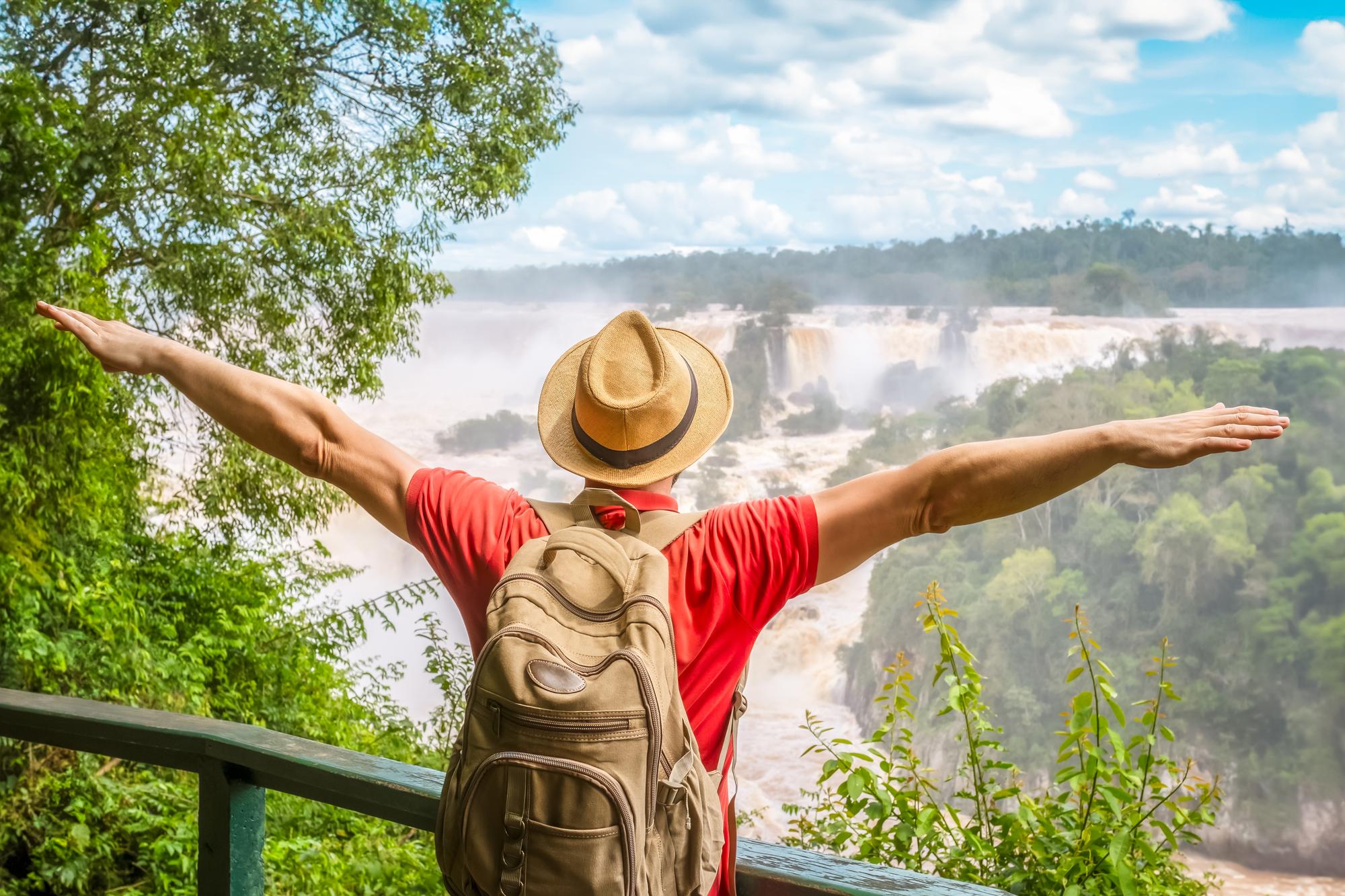 Turista de chapéu de palha, camiseta vermelha e mochila, de costas e de braços abertos no mirante, diante das quedas das Cataratas do Iguaçu cercadas de mata, em Foz do Iguaçu.