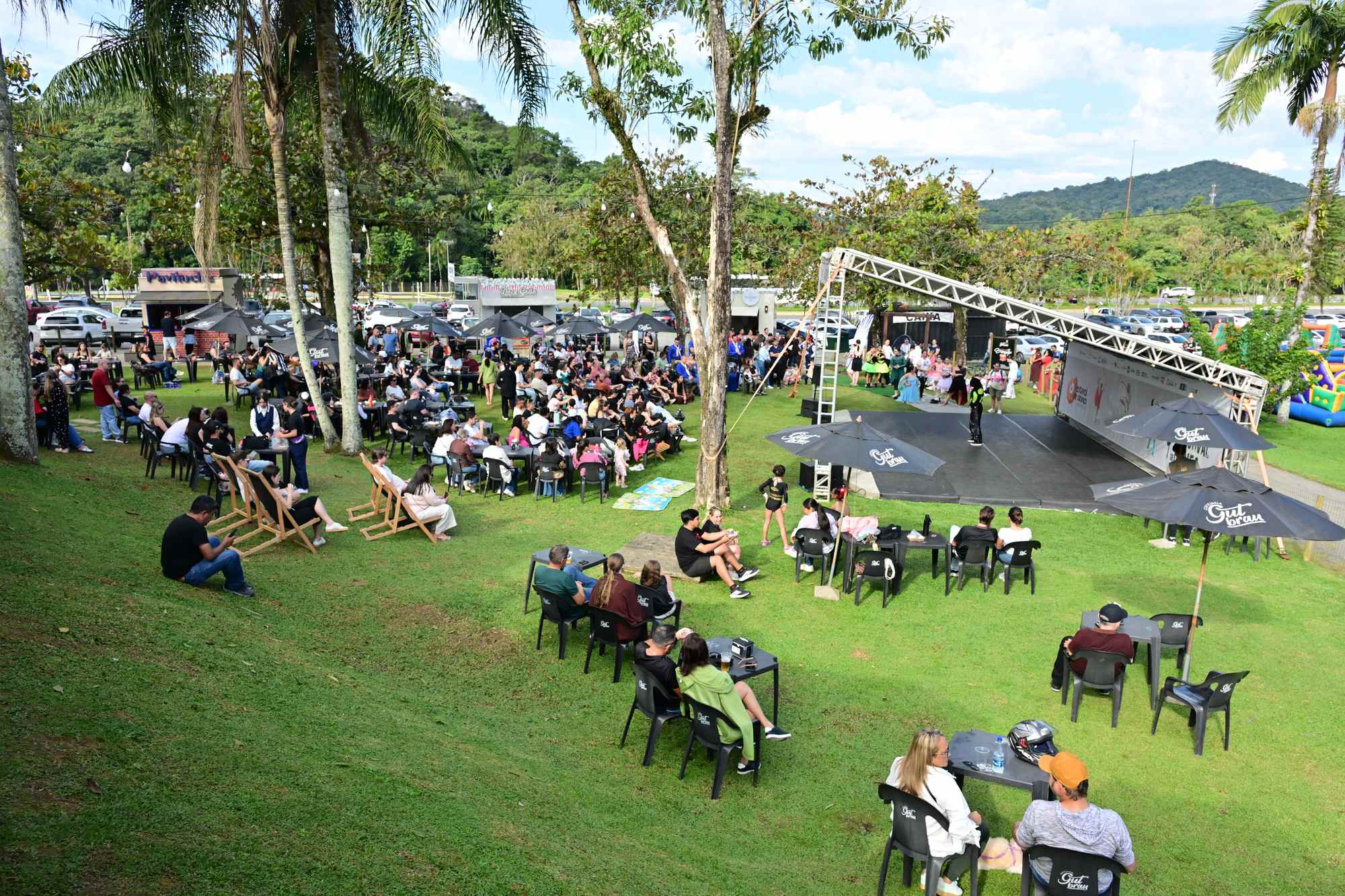 Público sentado em mesas e cadeiras de praia num gramado em Joinville, assistindo a uma apresentação num palco aberto do Festival de Dança, com palmeiras e os morros verdes da Serra do Mar ao fundo, num dia de sol.