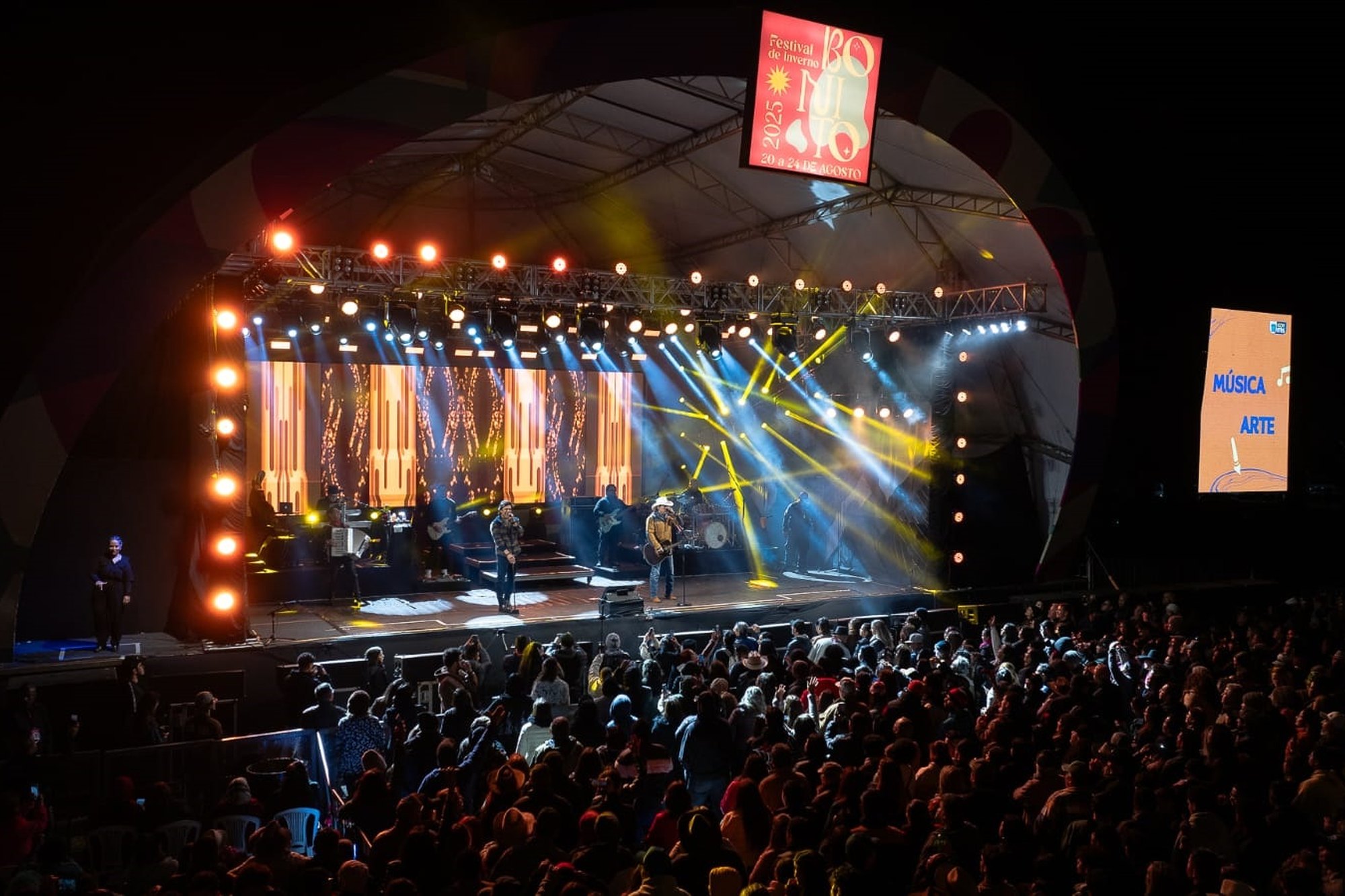 Palco principal do Festival de Inverno de Bonito iluminado à noite, visto do alto, com a plateia lotada em frente e telões nas laterais.