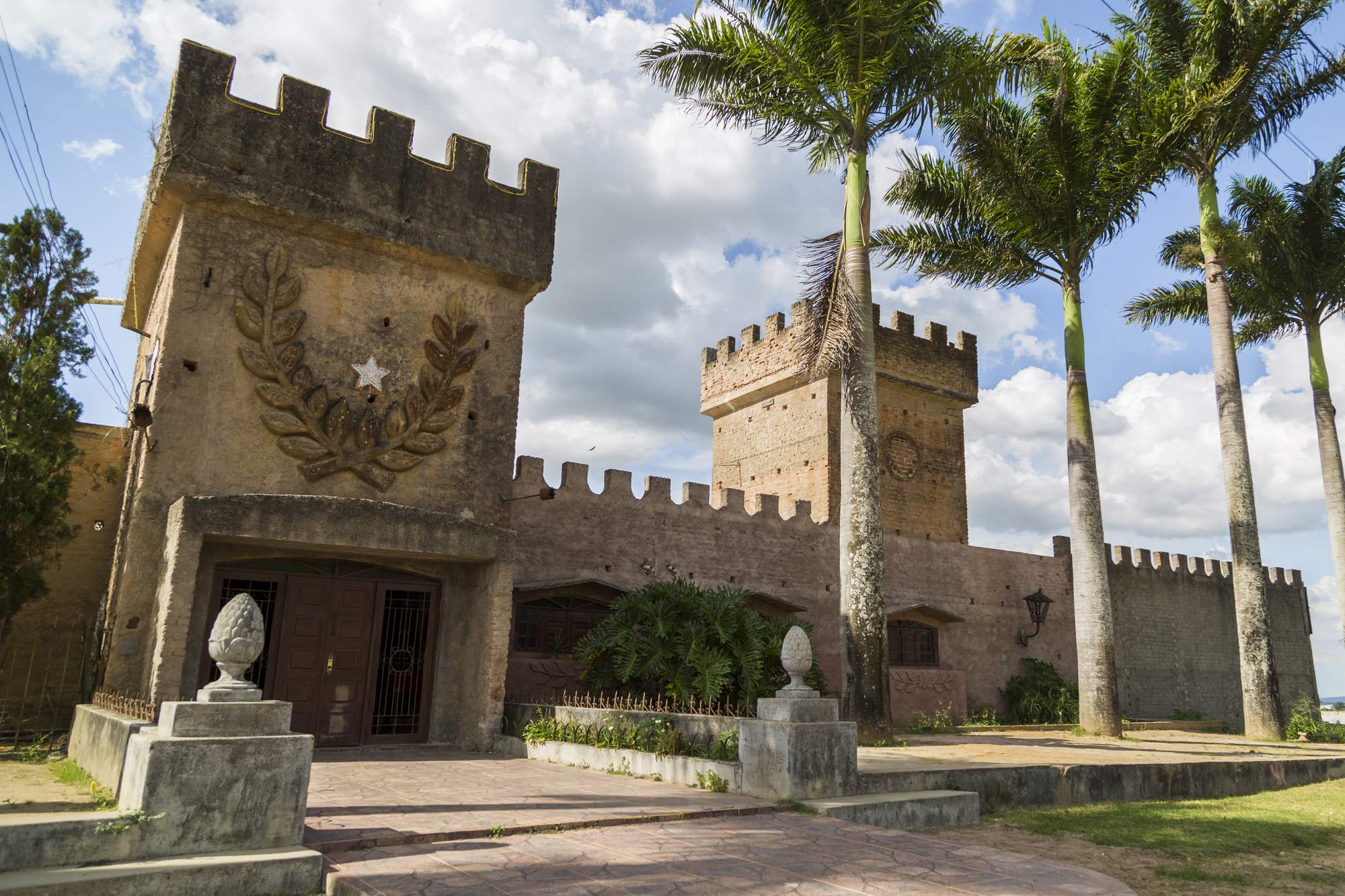 Castelo de João Capão, em Garanhuns, com torres de pedra com ameias, um brasão na fachada e palmeiras imperiais altas diante do céu azul com nuvens.