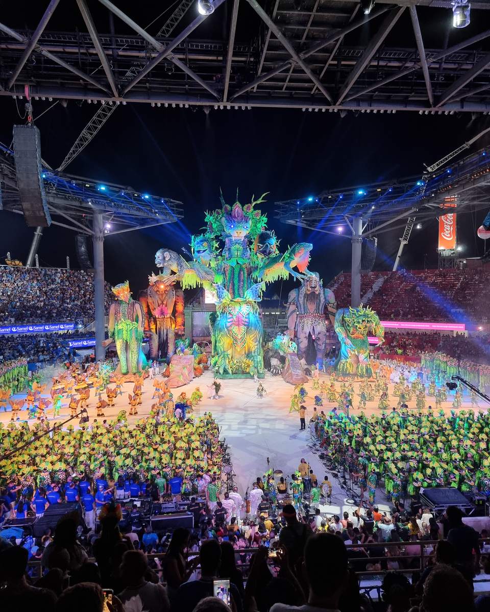 Arquibancada cheia no Bumbódromo de Parintins durante a apresentação, com o público da frente filmando a arena pelo celular e alegorias iluminadas ao fundo.