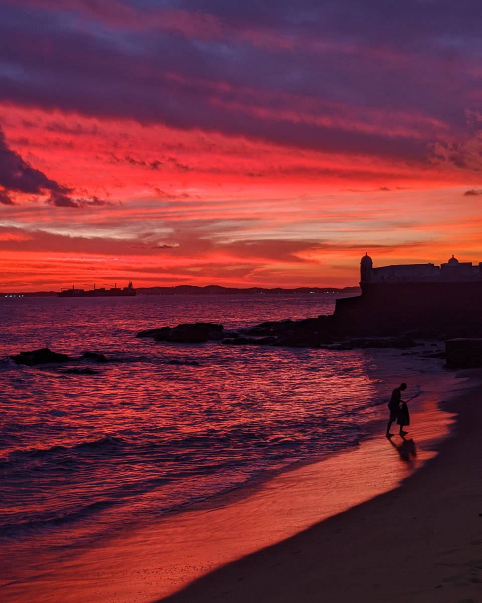 Pôr do sol vermelho e roxo sobre a Baía de Todos-os-Santos, visto da areia do Porto da Barra, em Salvador, com a silhueta do Forte de Santa Maria à direita e uma pessoa caminhando na beira da água.