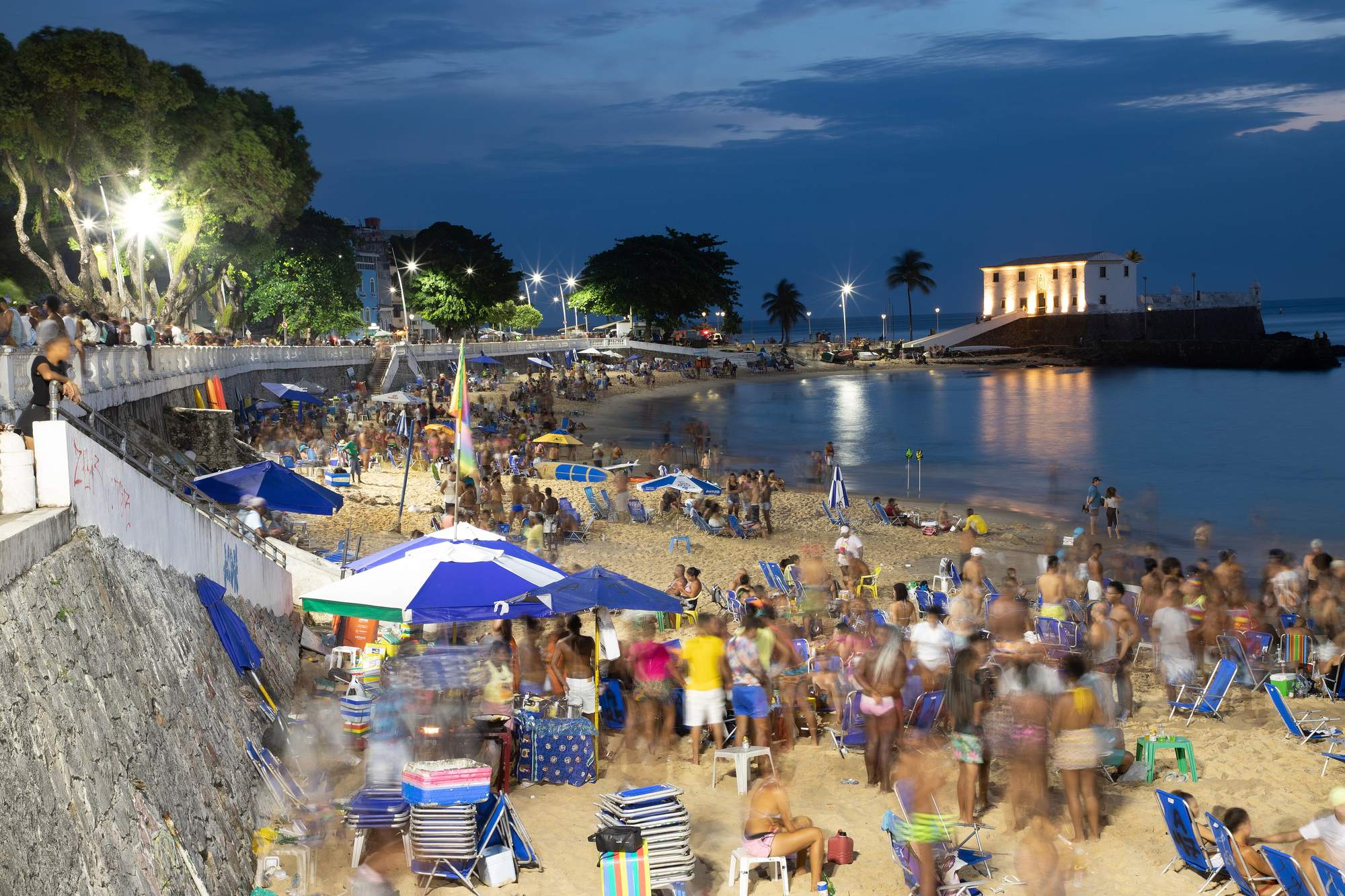 Praia do Porto da Barra, em Salvador, cheia de gente ao anoitecer, com guarda-sóis azuis e brancos, cadeiras de praia na areia, a orla iluminada e o Forte de Santa Maria aceso à beira do mar.