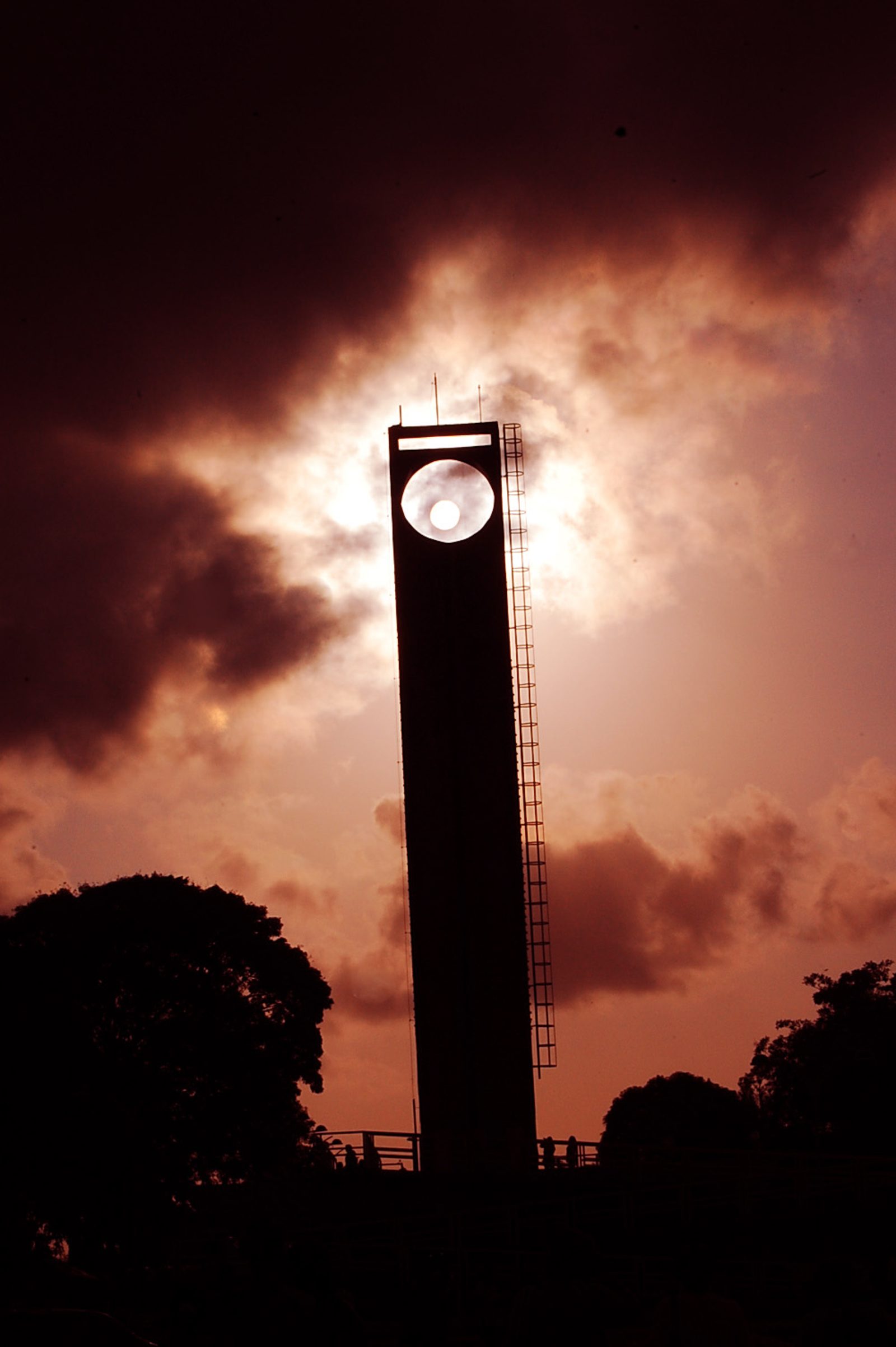 Silhueta do obelisco do Marco Zero do Equador contra o céu em tons alaranjados ao entardecer, com o sol visível bem no centro da abertura circular no topo do monumento.