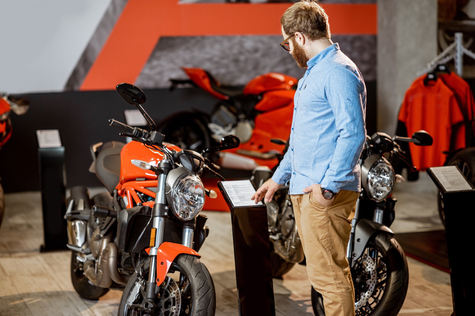 Homem de camisa azul e óculos lendo a ficha técnica de uma moto naked vermelha numa loja de motos, com outras motos esportivas e jaquetas expostas ao fundo.