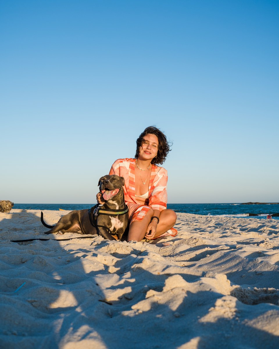 Jovem de cabelo cacheado sentada na areia da praia de Saquarema, no estado do Rio, ao lado de um pit bull cinza de peitoral e guia, no fim da tarde com o mar ao fundo.