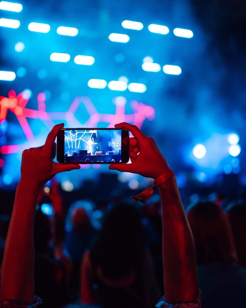Mãos erguidas segurando um celular na horizontal, filmando um show à noite, com o palco iluminado de azul e vermelho e a plateia de costas embaixo.