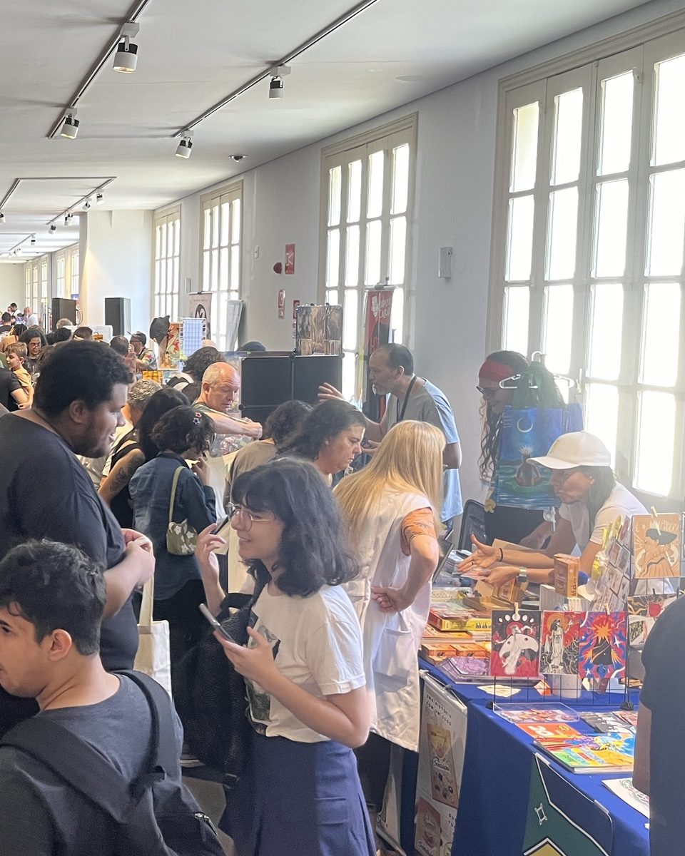 Visitantes circulando entre as mesas de artistas no FIQ BH 2024, no Minascentro, com uma jovem de óculos olhando o celular em primeiro plano e quadrinhos expostos sobre a toalha azul.