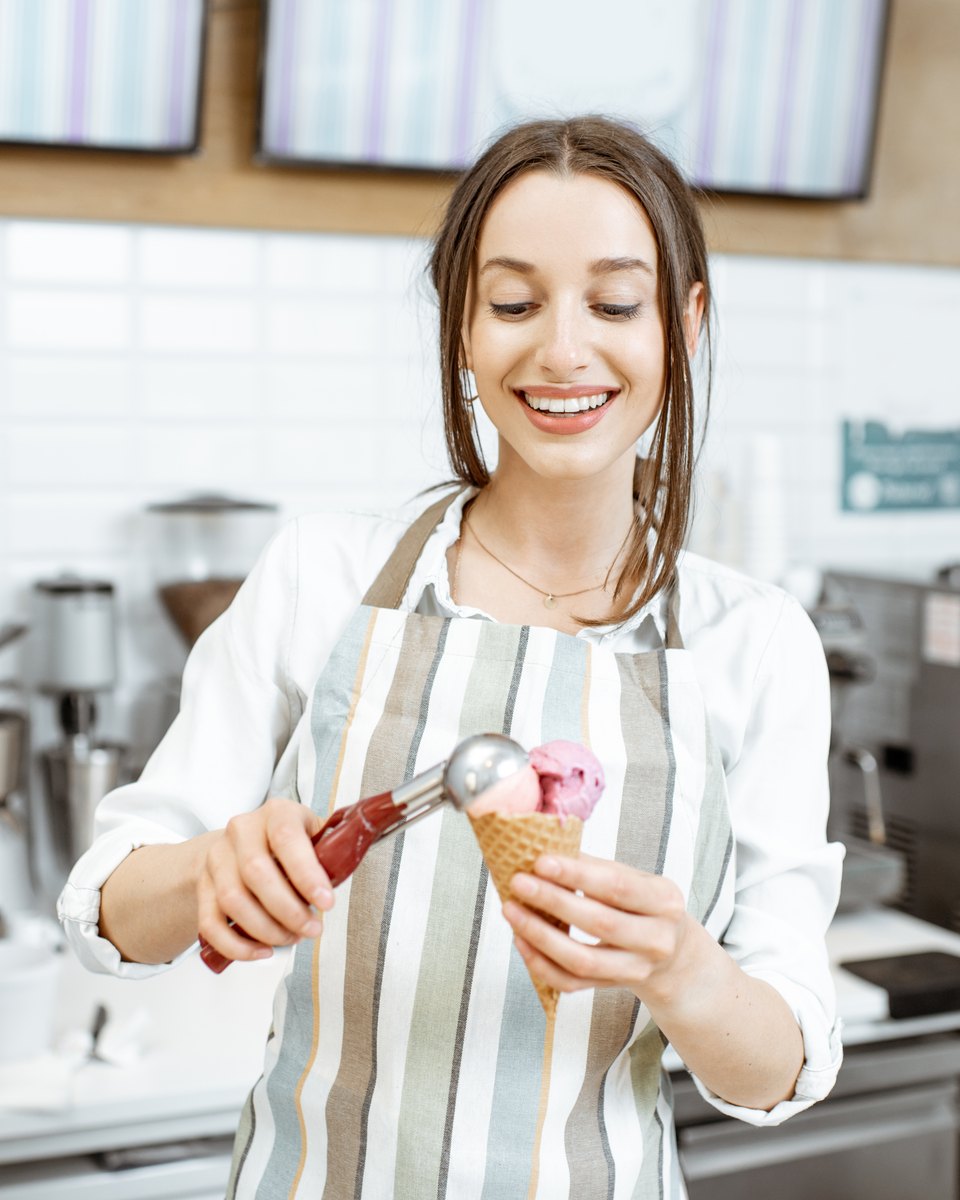 Atendente sorridente de avental listrado servindo uma bola de sorvete de frutas vermelhas numa casquinha, no balcão de uma sorveteria com máquinas de inox ao fundo.