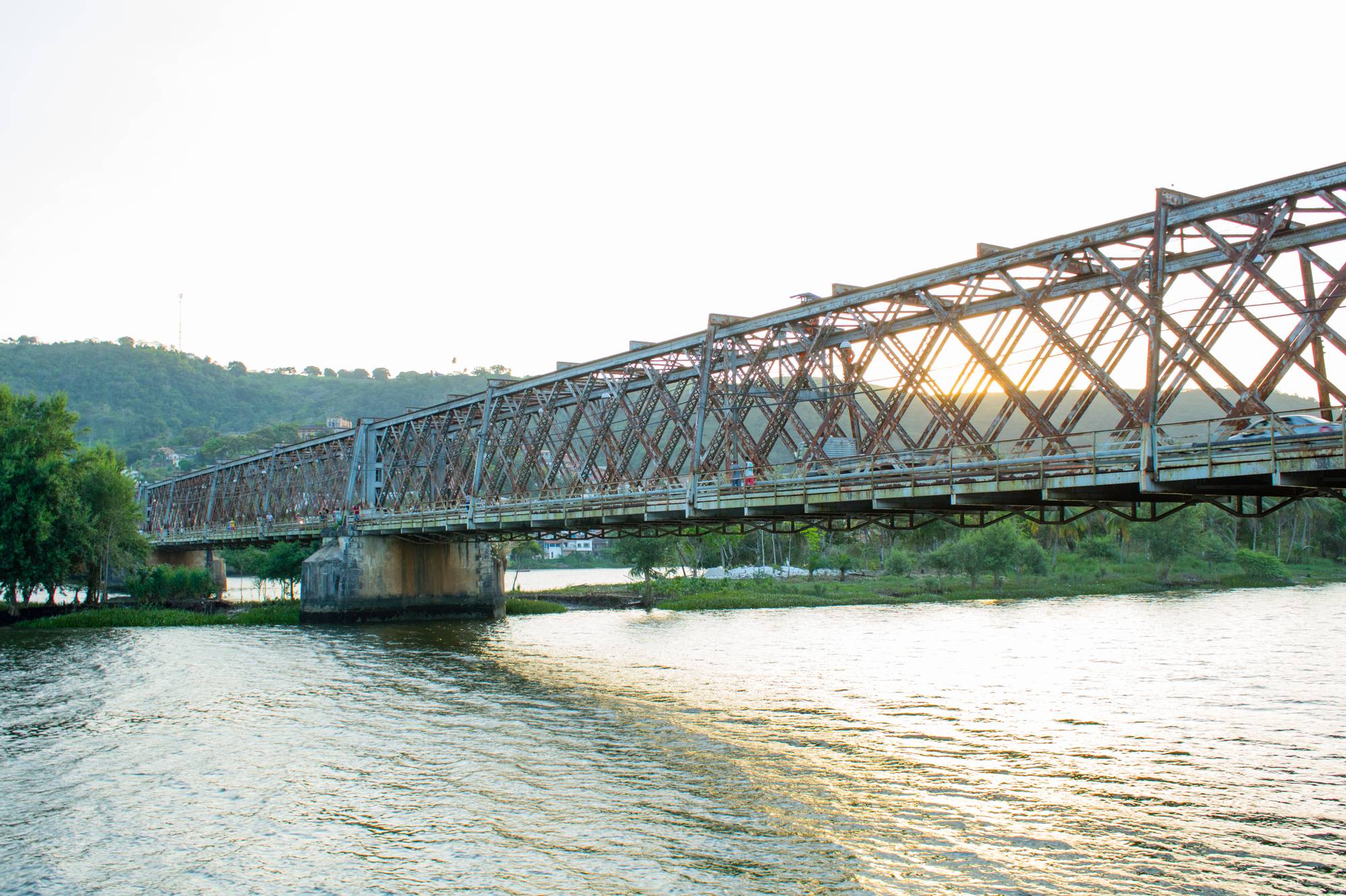 Ponte Dom Pedro II, de estrutura de ferro, atravessando o Rio Paraguaçu entre Cachoeira e São Félix, na Bahia, com a luz do sol passando pelas treliças e morros verdes ao fundo.