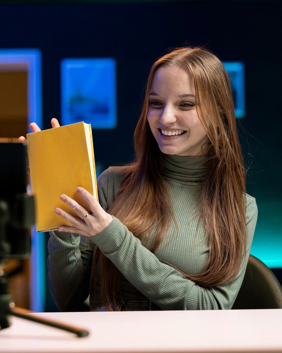 Jovem de cabelo longo e blusa verde sorrindo e mostrando um livro de capa amarela para um celular preso num tripé sobre a mesa, gravando uma resenha num quarto com luz azulada.