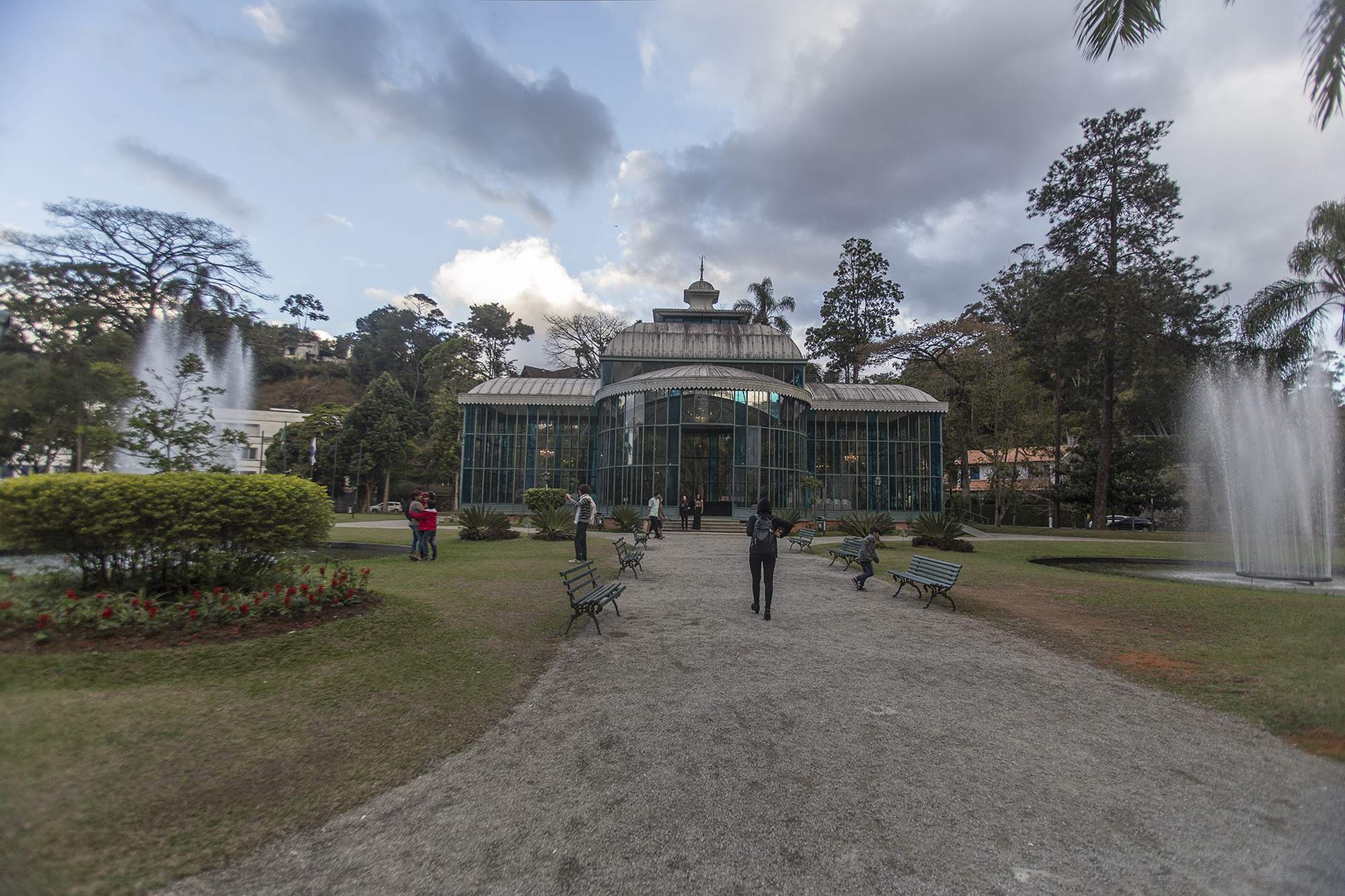 Visitantes caminhando e fotografando no jardim do Palácio de Cristal, em Petrópolis, com bancos de ferro verdes, canteiro de flores vermelhas, um chafariz à direita e o pavilhão de vidro e ferro ao fundo.
