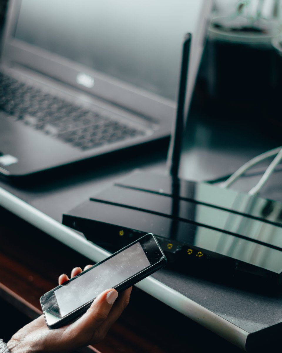 Mão segurando um celular ao lado de um roteador Wi-Fi de antenas, sobre uma mesa com notebook e vasos de plantas ao fundo.