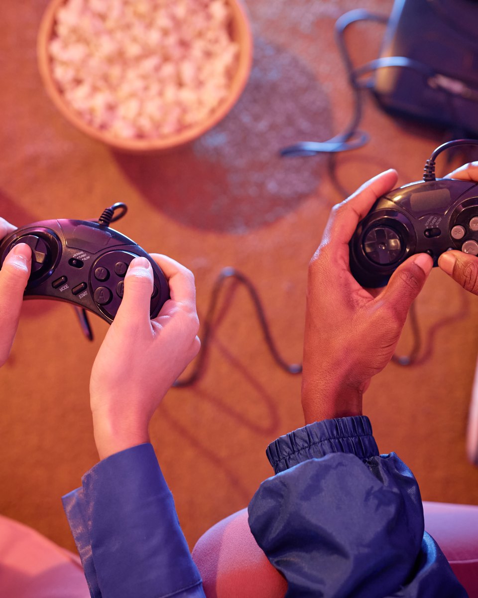 Vista de cima das mãos de duas pessoas segurando controles de videogame retrô pretos, sentadas no chão com um console antigo, um copo e uma tigela de pipoca.