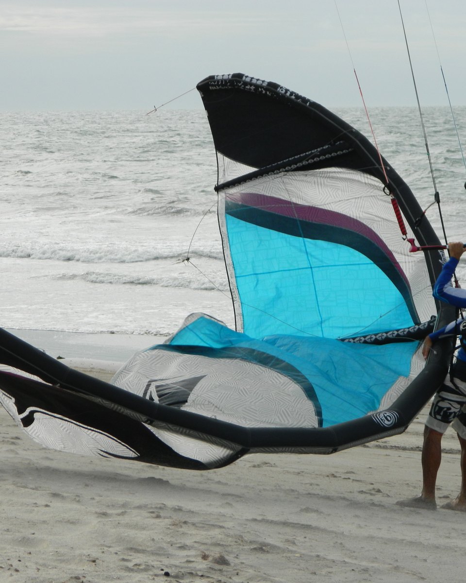 Kitesurfista de lycra azul segurando a barra de uma pipa branca, preta e azul-turquesa ainda na areia, preparando a decolagem numa praia de Jericoacoara, no Ceará, com o mar agitado ao fundo.