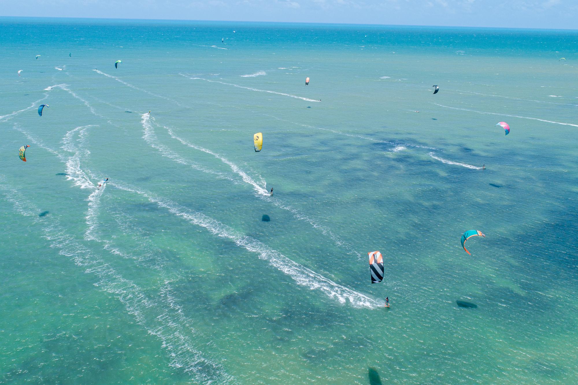 Vista aérea de dezenas de kitesurfistas deslizando sobre o mar verde-azulado do litoral oeste do Ceará, em Cumbuco, deixando rastros de espuma branca na água.