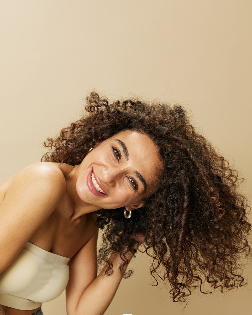 Mulher sorridente de cabelo cacheado volumoso inclinando a cabeça para o lado, contra um fundo bege.