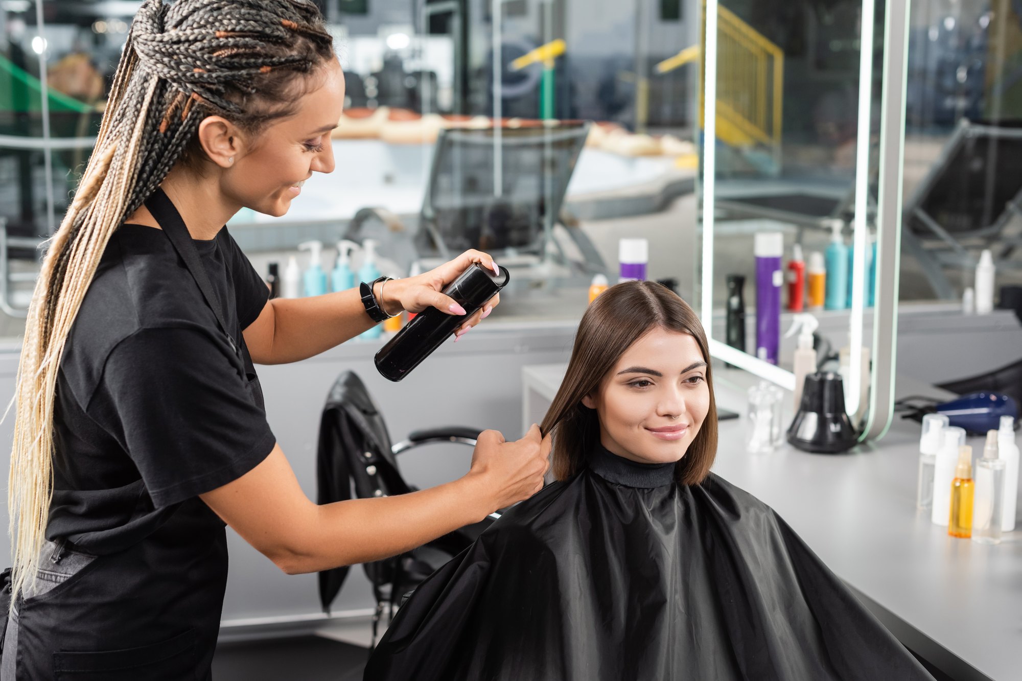 Cabeleireira de tranças longas finalizando com spray o cabelo liso de uma cliente morena, sentada na cadeira de um salão com produtos na bancada.