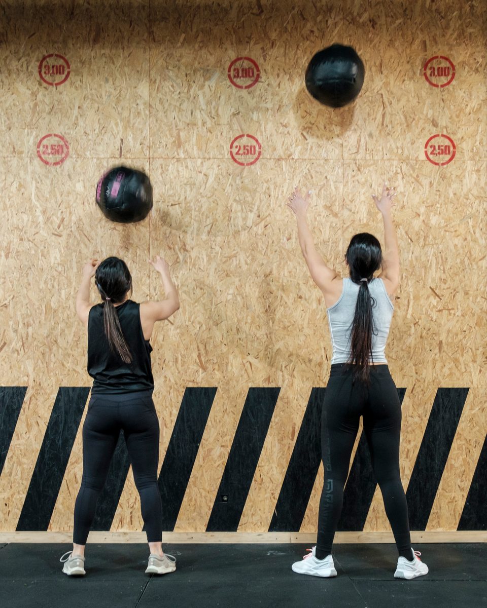 Duas mulheres de costas, de legging preta e cabelo preso, arremessando bolas de peso contra uma parede de madeira com alvos marcados, numa academia de treino funcional.