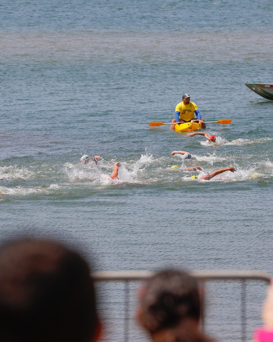 Nadadores de touca no Lago Paranoá, em Brasília, acompanhados por um caiaque e um barco de apoio, e o público de costas na grade da margem, com uma pessoa filmando a prova com o celular, na etapa de Brasília da Copa do Mundo de Triathlon de 2023.