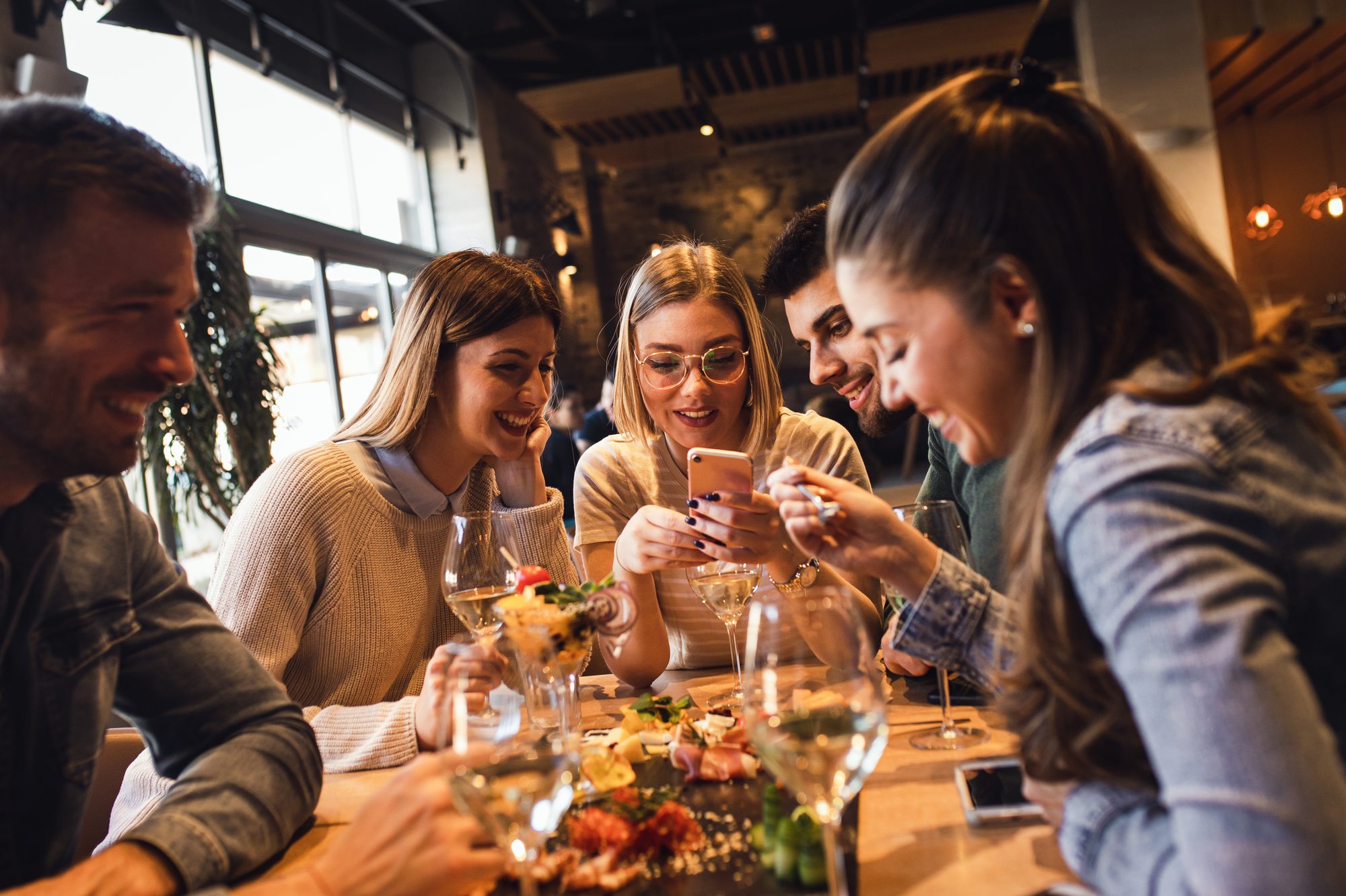 Cinco amigos rindo em volta de uma mesa de restaurante com petiscos e taças de vinho branco, enquanto uma jovem de óculos segura o celular.