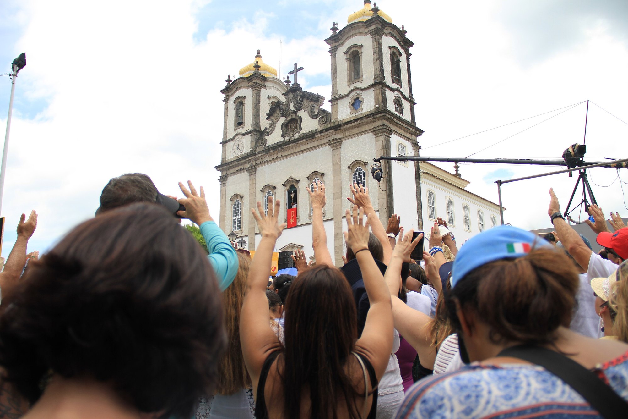 Multidão de costas, de braços erguidos, diante da Basílica do Senhor do Bonfim, em Salvador, durante a Lavagem, com fiéis filmando com o celular e as duas torres brancas da igreja ao fundo.