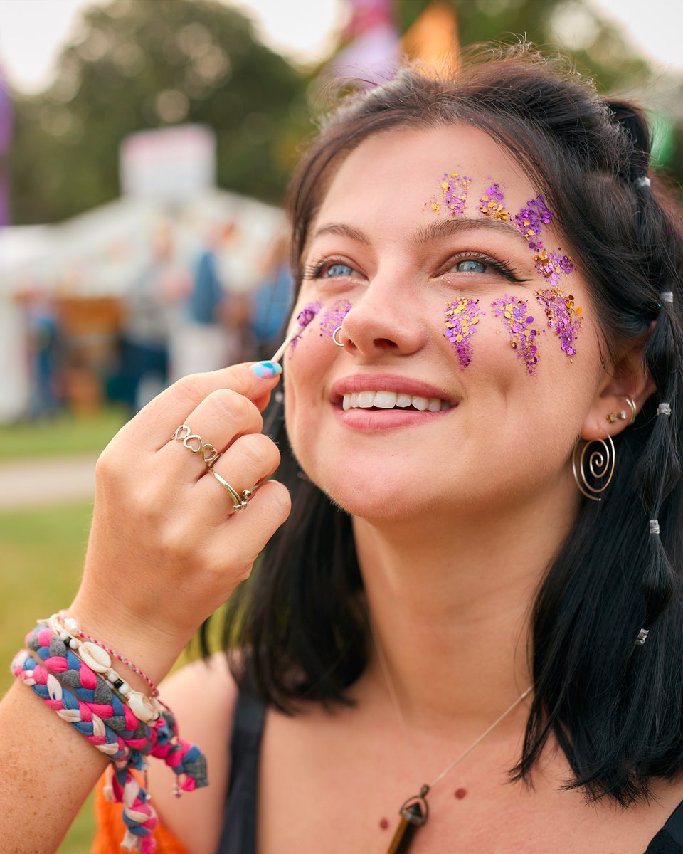 Jovem sorrindo enquanto uma amiga aplica glitter roxo e dourado no rosto dela, num festival de música ao ar livre com tendas ao fundo.