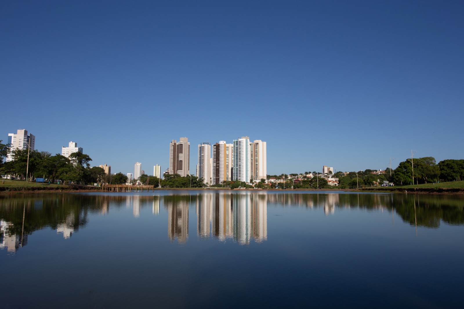 Lago do Parque das Nações Indígenas, em Campo Grande, numa manhã de céu limpo, com os prédios altos refletidos na água parada e a mata em volta.