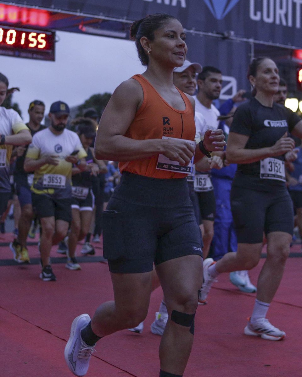 Corredora de regata laranja e outros atletas com número de peito saindo da largada da Maratona de Curitiba ainda no escuro, alguns olhando o relógio no pulso.