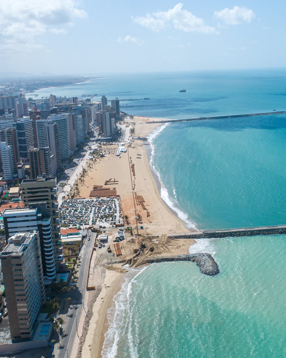 Vista aérea da Avenida Beira-Mar, em Fortaleza, com os prédios da orla, a faixa de areia do aterro, os espigões de pedra entrando no mar verde e o píer ao fundo.