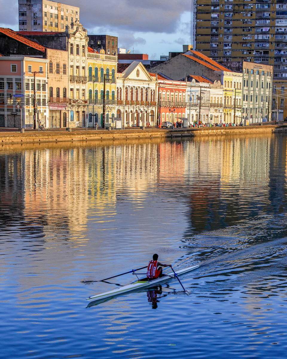 Remador de regata vermelha num barco de competição no Rio Capibaribe, no Recife, ao fim da tarde, com os casarões coloridos da Rua da Aurora refletidos na água.